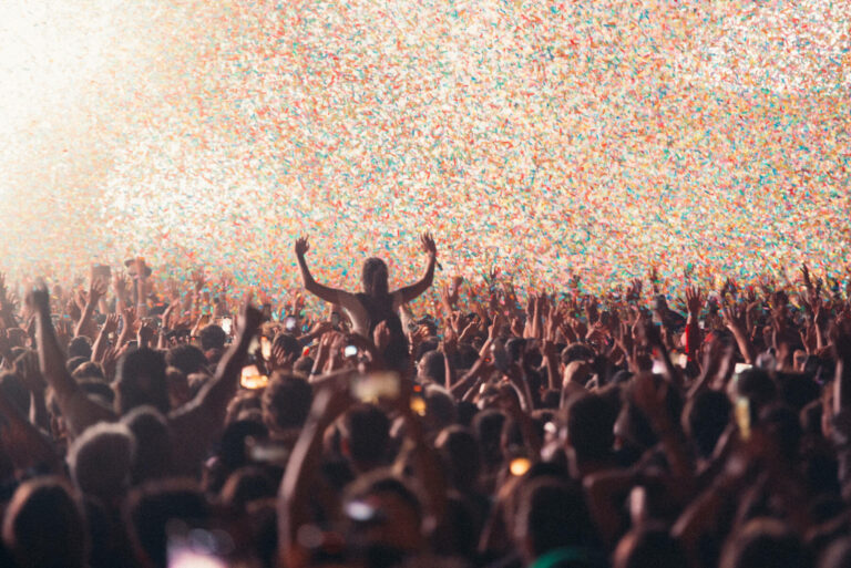 A crowd of people at a concert with their hands raised, enjoying a confetti explosion.