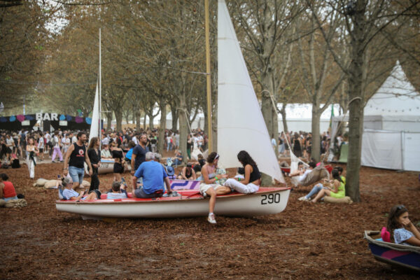 An outdoor festival scene with people relaxing and socializing in and around sailboats placed on the ground covered with brown leaves. There are trees in the background and a sign for a bar.