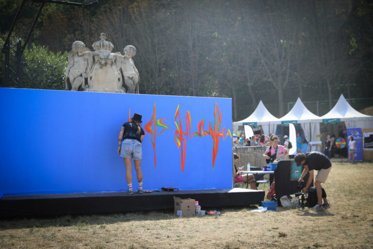 A person is painting orange and yellow graffiti on a large blue wall at an outdoor event with tents and bystanders in the background, with a statue atop a structure visible behind the wall.