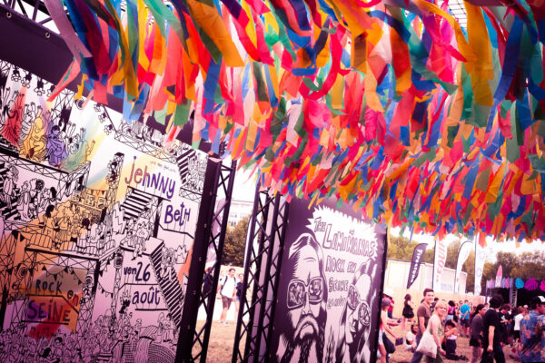A vibrant festival scene with colorful streamers hanging overhead and people milling about. Billboards with comic-style illustrations and text announce performers for an event dated August 26.