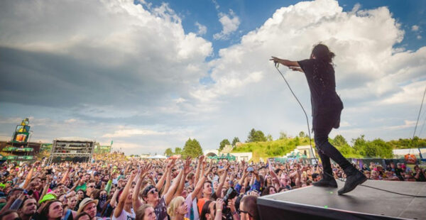 A performer on stage at an outdoor concert gesturing to an enthusiastic crowd with hands raised under a blue sky with clouds.