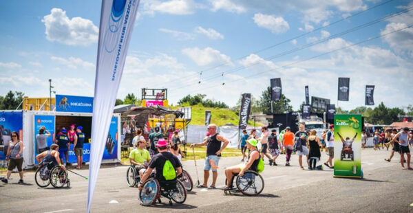 Alt text: A sunny day at an outdoor event with people walking and several individuals using handcycles. Flags and banners of different brands are displayed around the area, with booths and festival structures in the background.