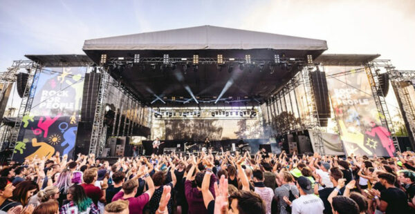 Outdoor music festival with a crowd of people raising their hands towards a stage with large banners reading 