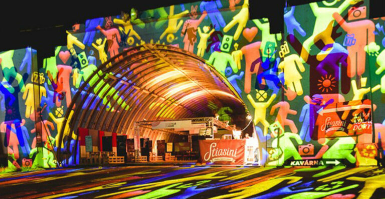 Colorful light projections of abstract human figures and symbols on the exterior of a curved building structure at night.