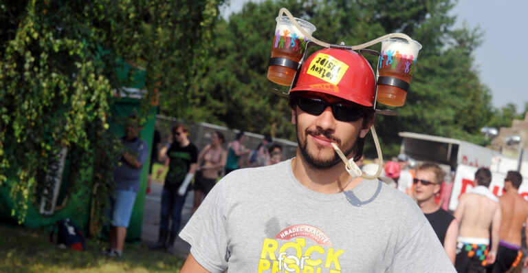 A man wearing a beer helmet with two cups attached walks at an outdoor event, with people in the background.