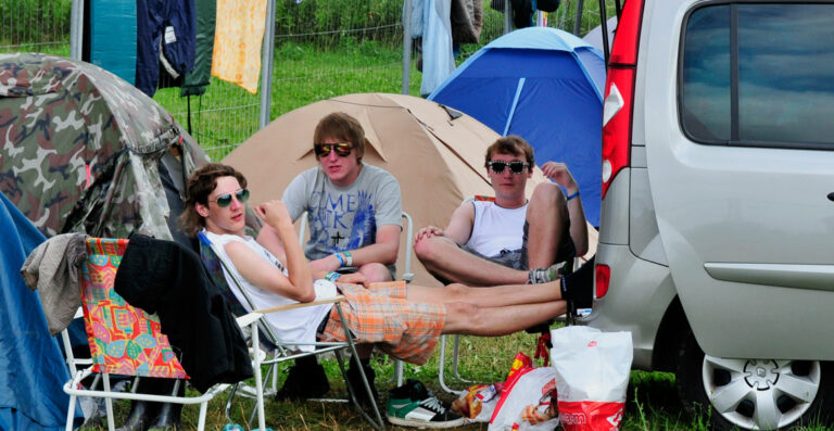 Three young individuals are sitting in camping chairs outside a tent beside a parked car at a campsite, with various camping items strewn around them.