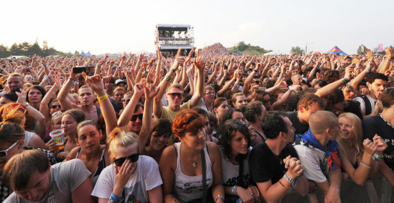 A large crowd at an outdoor concert raising their hands in the air with enthusiasm.