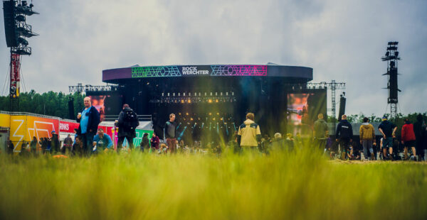 Concert audience in a grassy field with a view of a large stage featuring the words 