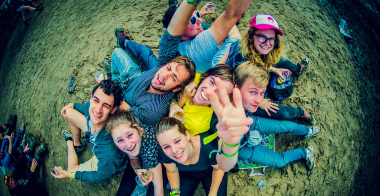 A group of young adults lying on the ground in a circle, smiling and making peace signs, at an outdoor event with a festival atmosphere.