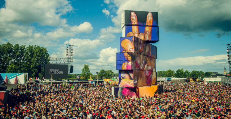 A large crowd gathered at an outdoor music festival with a towering sculpture of a hand making a peace sign, beneath a blue sky with clouds.