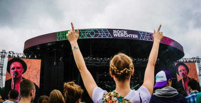 Concert audience with raised hands at the Rock Werchter festival, with large screen displaying a performer and stage in the background.
