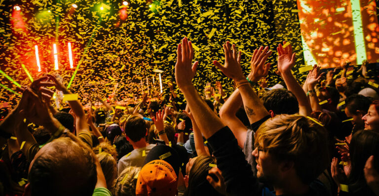 A crowd of people at a concert with raised hands, celebrating as confetti falls around them, with stage lights visible in the background.