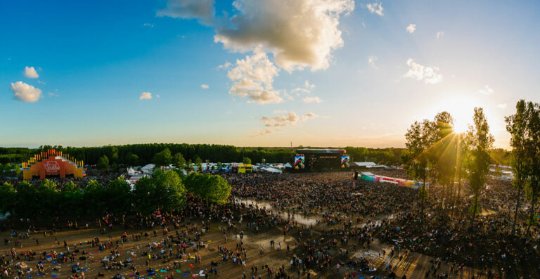 Panoramic view of an outdoor music festival at sunset with large crowds, a stage in the distance, and the sun low in the sky amidst trees.