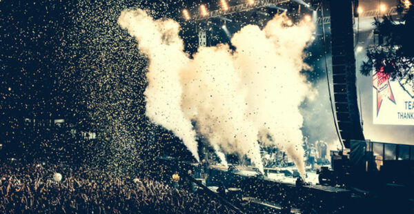 A large crowd at a music festival with confetti in the air and plumes of smoke on stage under a banner that says 