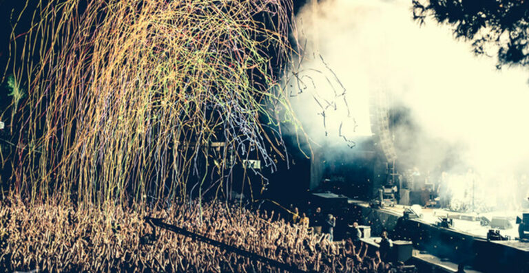 A concert scene with streams of colorful confetti raining down on a cheering crowd with stage lights and smoke in the background.