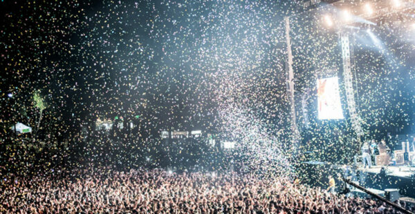 A crowd at a concert with confetti falling from above, backlit by stage lighting with a screen showing a performer.