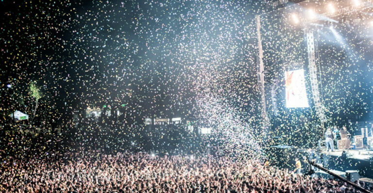 A crowd at a concert with confetti falling from above, backlit by stage lighting with a screen showing a performer.