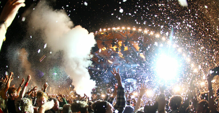A vibrant concert scene with a crowd cheering and throwing their hands up in the air under a shower of confetti, with stage lights shining brightly from the center.