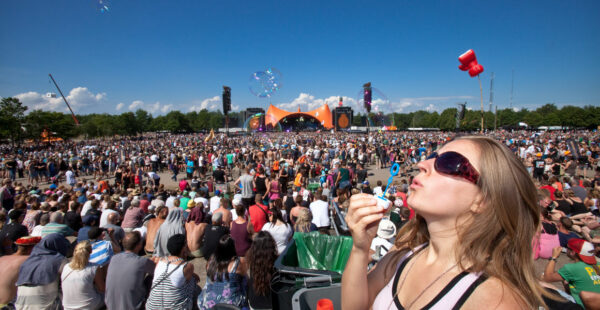 A woman blowing bubbles in the foreground with sunglasses on, at a crowded outdoor festival with a large stage in the background under a clear blue sky.