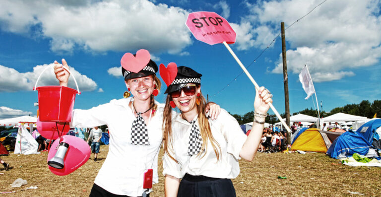 Two cheerful people wearing heart-shaped headbands and checkered hats are posing at a festival with a red bucket and a sign that says 