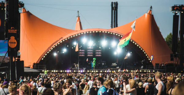 A large outdoor music festival crowd during daytime with people watching a performance under a big orange tented stage, some holding flags, with stage lights visible.