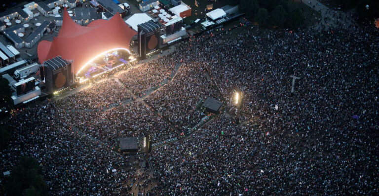 An aerial view of a densely crowded outdoor music festival at dusk featuring a large stage with bright lights and a red canopy.