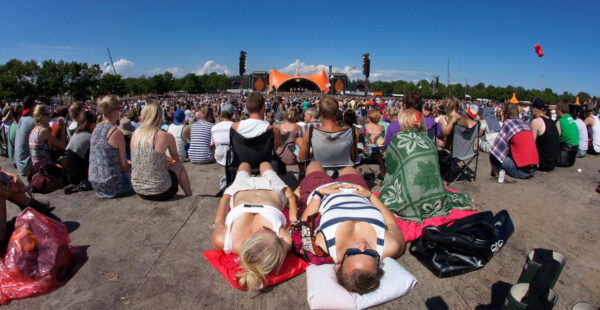 A crowded outdoor festival with people sitting on the ground and two individuals lying in the foreground, facing an orange-hued stage under a blue sky.