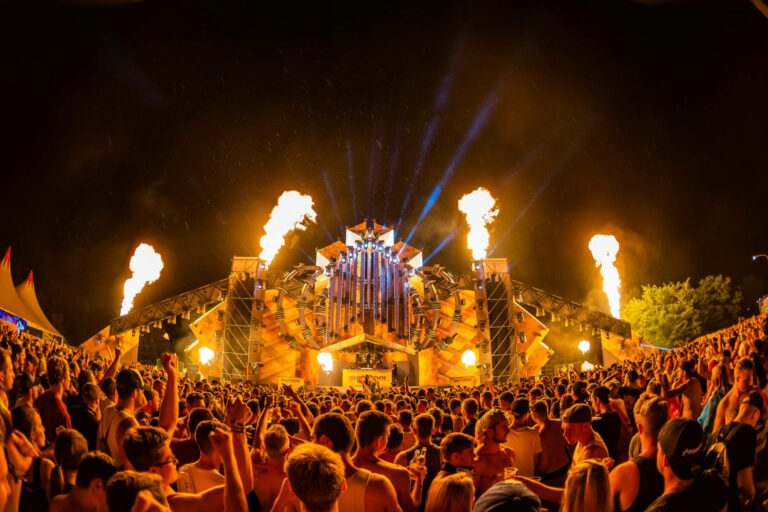 A music festival at night with a large crowd facing an elaborate stage that features pyrotechnics and dramatic lighting.