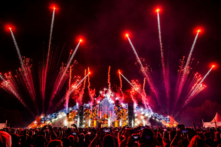 A vibrant outdoor music festival at night with a crowd of spectators, a stage with elaborate lighting, and an impressive fireworks display.