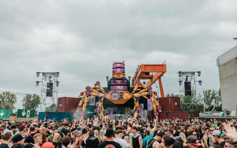 A large crowd of people enjoying an outdoor music festival with a colorful, robot-like stage decoration in the background.