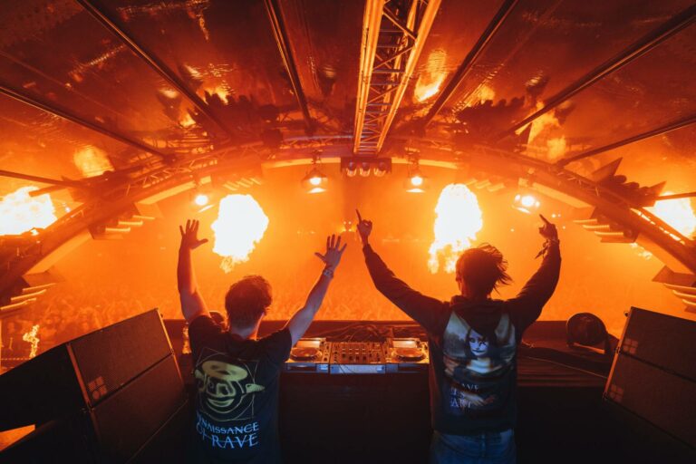 Two DJs with raised arms are performing in front of a lively crowd at a music event, with intense pyrotechnics creating large flames on stage.