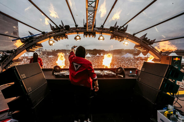 A DJ facing a large crowd from a stage with turntables, raising their hand up in the air with bursts of flame shooting up from the stage against a cloudy sky.