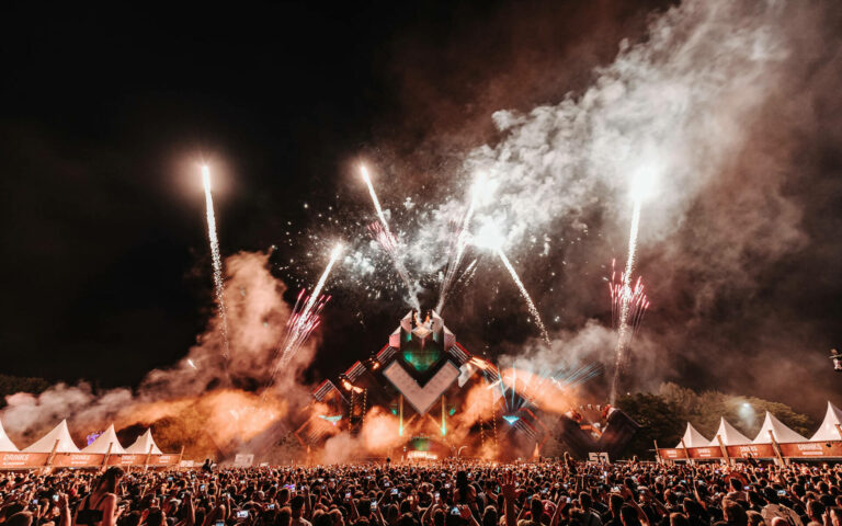 A large crowd at an outdoor music festival at night, with a central stage illuminated by fireworks and stage lights.