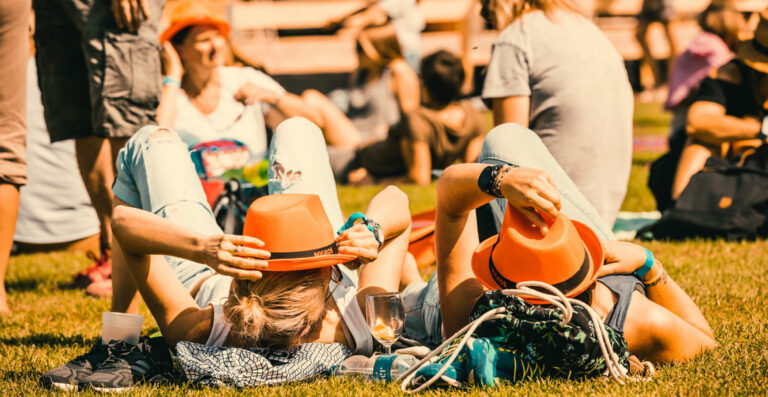 People relaxing on a sunny day at a park, with two individuals in the foreground lying down on the grass with their hats over their faces.