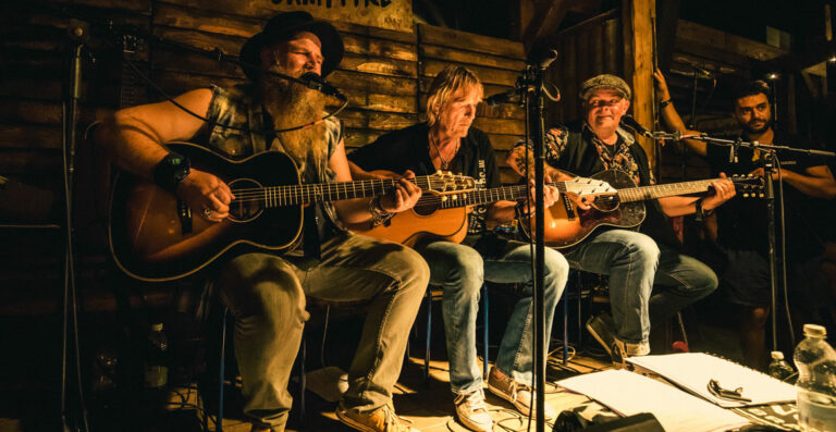 Four musicians seated on stage performing an acoustic set, with the focus on two guitarists in the center, illuminated by warm stage lighting in a rustic wooden venue.