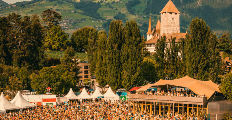 A crowded outdoor festival with tents in the foreground and a historic castle with a tower in the background, set against a scenic backdrop of green hills and trees.