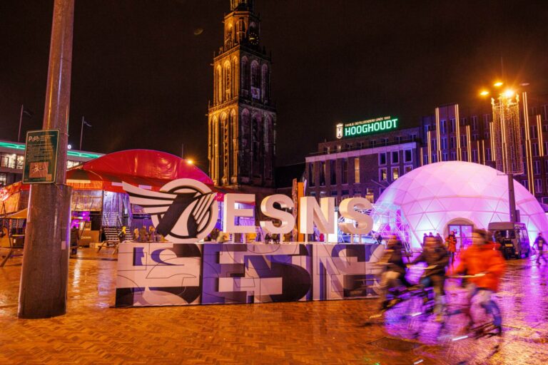 A vibrant night scene with large block letters spelling "SENS" in the foreground. Behind the text, there's a domed structure illuminated with purple and blue lights, and cyclists passing by in a blur. In the background, a historic tower stands illuminated, and a sign with "HOOGHOUDT" is visible above a building. The sky is dark, and a small crescent moon is visible.