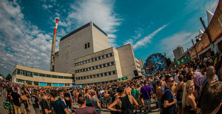 Outdoor music festival with a large crowd of people in front of an industrial setting with a tall chimney and buildings under a blue sky with fluffy clouds.