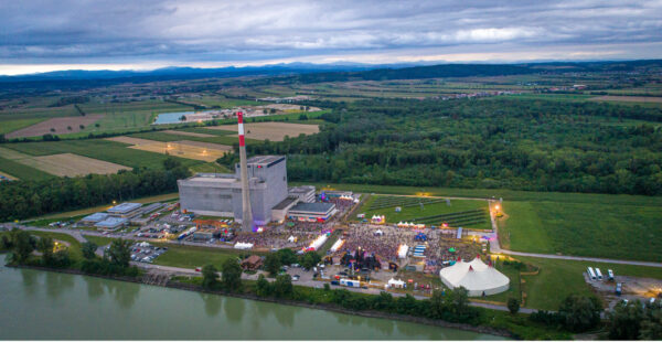 Aerial view of a large outdoor event near a river with a concrete industrial structure featuring a tall red-and-white smokestack, surrounded by agricultural fields and distant hills under a cloudy sky.