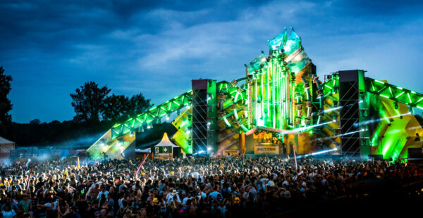 A large crowd of people at an outdoor music festival in the evening, with a brightly lit stage featuring green lights and an elaborate set design reminiscent of a futuristic structure.