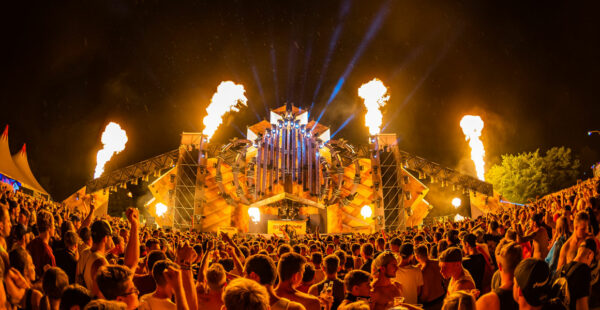 A vibrant outdoor music festival at night with a large crowd of people facing a stage that features an elaborate set design with lights and pyrotechnic flames shooting into the air.