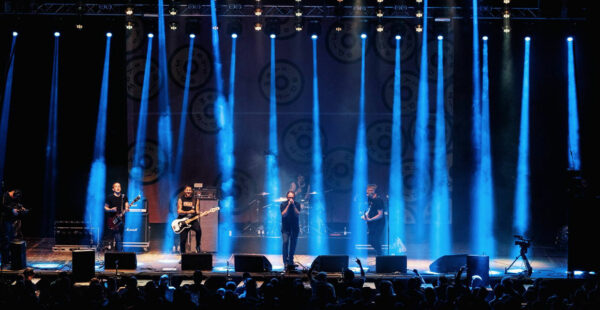A band performing on stage with blue stage lighting, audience in the foreground, and banners with logos in the background.