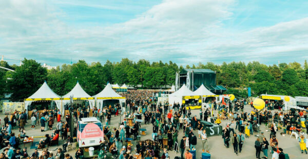 Outdoor festival scene with a crowd of people, food tents, and a stage set up in a park-like setting with trees and a clear sky.