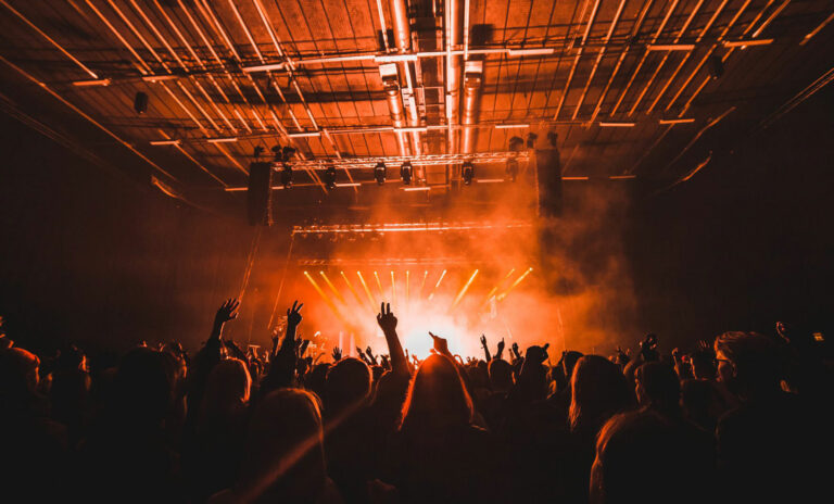 Silhouetted concert crowd with raised hands against a brightly lit stage with beams of light and haze.