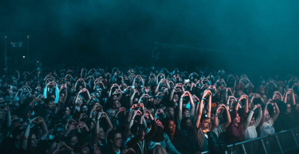 A crowd of concert-goers with many people forming heart shapes with their hands, illuminated by stage lights in a dimly lit venue.
