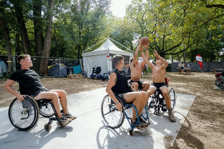 A group of young men playing wheelchair basketball outdoors, with two of them reaching for the ball in the air.