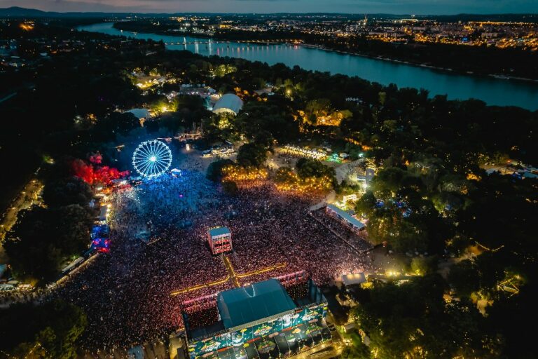 Aerial night view of a crowded festival with lights, a large Ferris wheel illuminated, and a river in the background.
