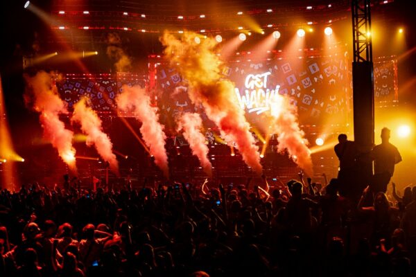A concert scene with intense stage lighting and pyrotechnics, a dense crowd of spectators with raised hands, and large text 