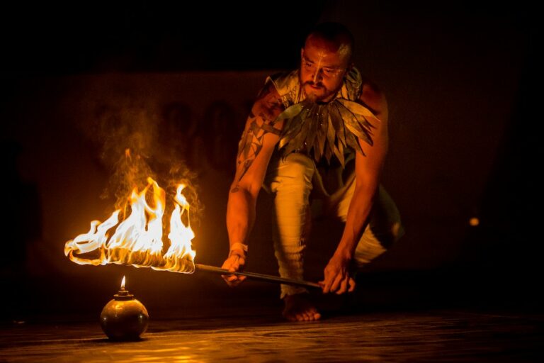 A performer with tattoos and leaf-like costume elements crouches by a flaming pot, holding a long staff with a fire-lit end, against a dark background with dimly visible spectators.