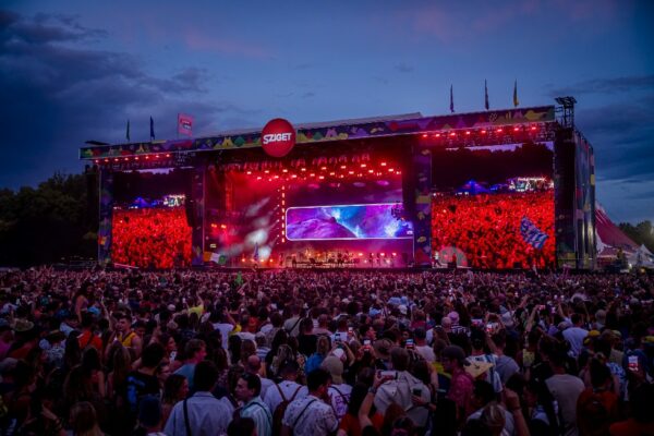 A large crowd of concertgoers enjoying an outdoor music festival at dusk, with a brightly lit stage and colorful light show in the background.